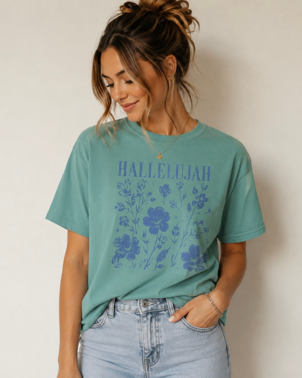Woman looking down wearing a teal Hallelujah floral Christian T-shirt with blue lettering against an off-white studio background.
