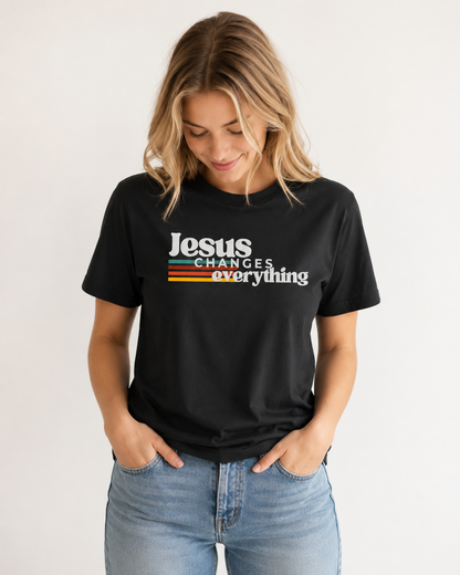 Blonde woman looking down with her hands in her pockets, wearing a black Jesus Changes Everything Christian T-shirt against an off-white studio background.