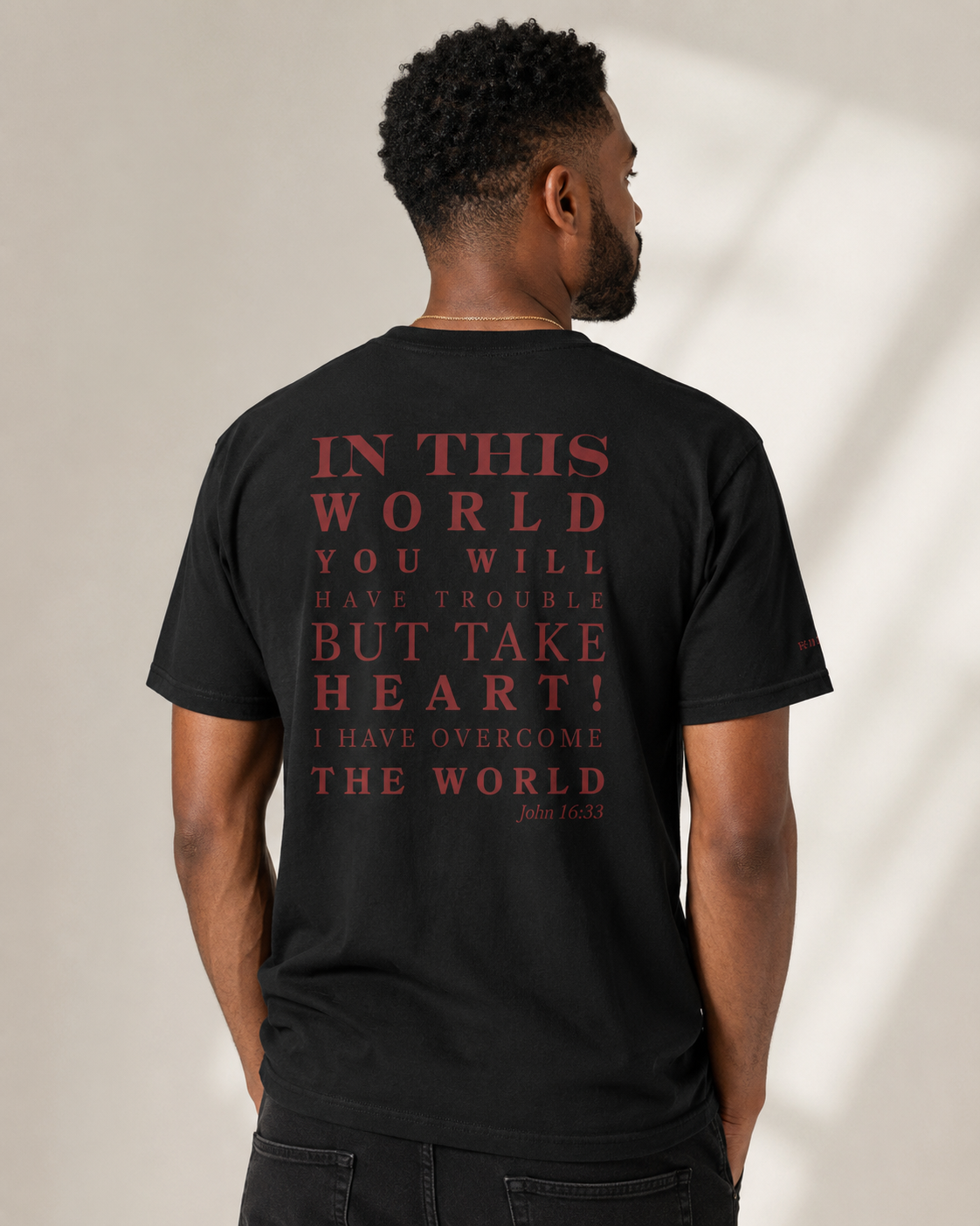 Male model wearing a black John 16:33 Christian t-shirt featuring the red Take Heart scripture graphic on the back.