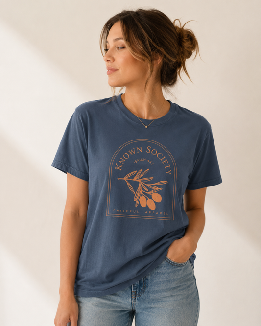 Woman modeling a blue Known Society Isaiah 43:1 Christian T-shirt against an off-white studio background.